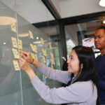 Female business analyst working with product owner using sticky notes on white glass wall